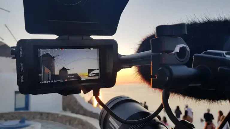 Eine Kamera von black@white media mit aufgeklappten Sucher vor den Windmühlen von Mykonos. Im Display sind 3 weiß gestrichene Windmühlen gestaffelt am Meer vor einem Sonnenuntergang zu sehen.